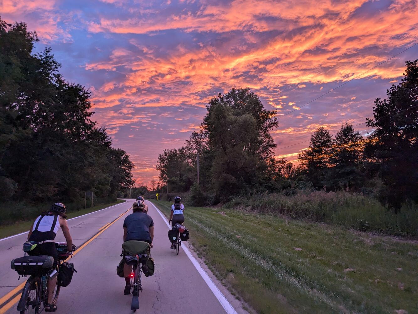 A group of cyclists riding down an empty road towards a colorful sunset