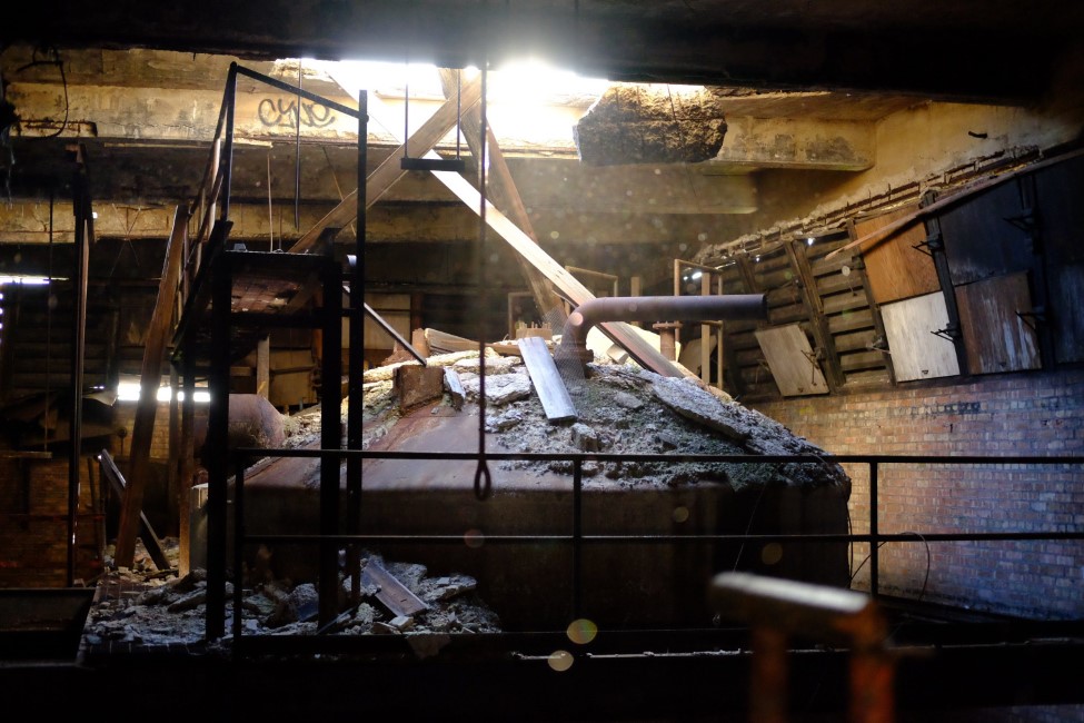 a large, rusting metal tank in a decaying building. the partially collapsed roof has dropped rubble on top of the tank, and allows light to stream into the otherwise dark room, making the dust particles in the air glow.
