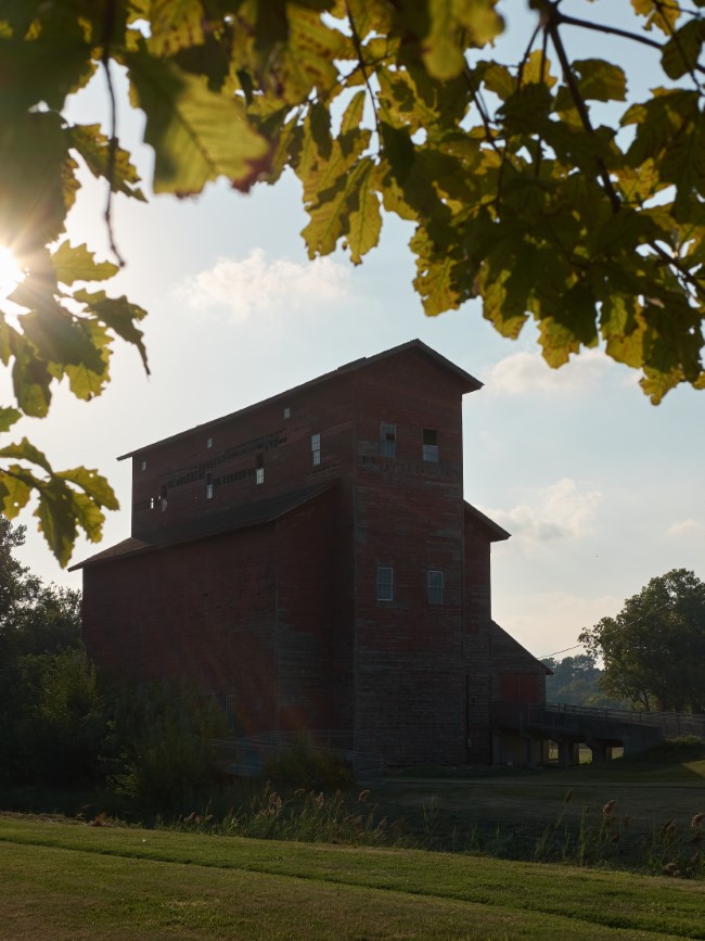 an old, wooden grain elevator, painted with weathered red paint. leaves in the foreground obscure the setting sun backlighting the scene.