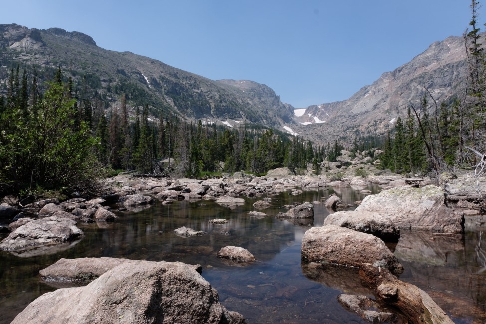 a secluded pond, filled with still water and large rocks, surrounded by trees and mountains.