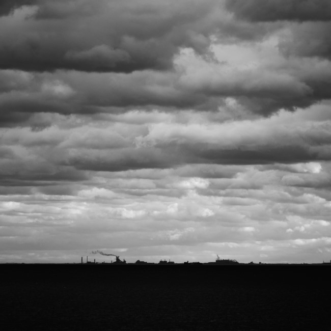 a monochrome image of factory buildings seen from across lake michigan. the distance makes the large buildings appear tiny compared to the water and sky.