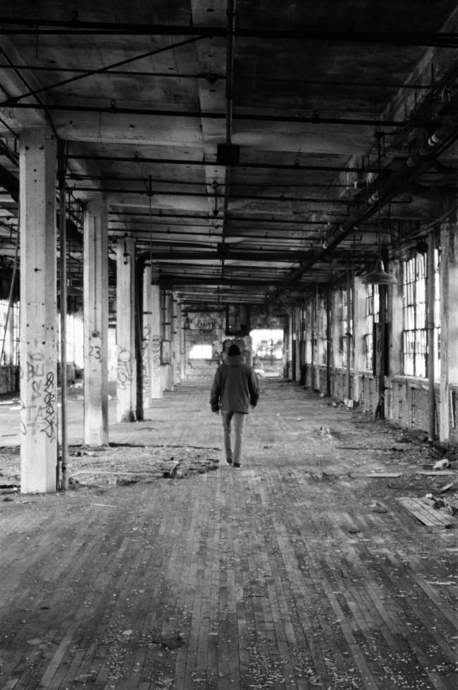a black and white photo of a man walking away from the camera, with his back turned to it. the room he is in is large, and empty save some graffitied concrete pillars and rubble on the ground.