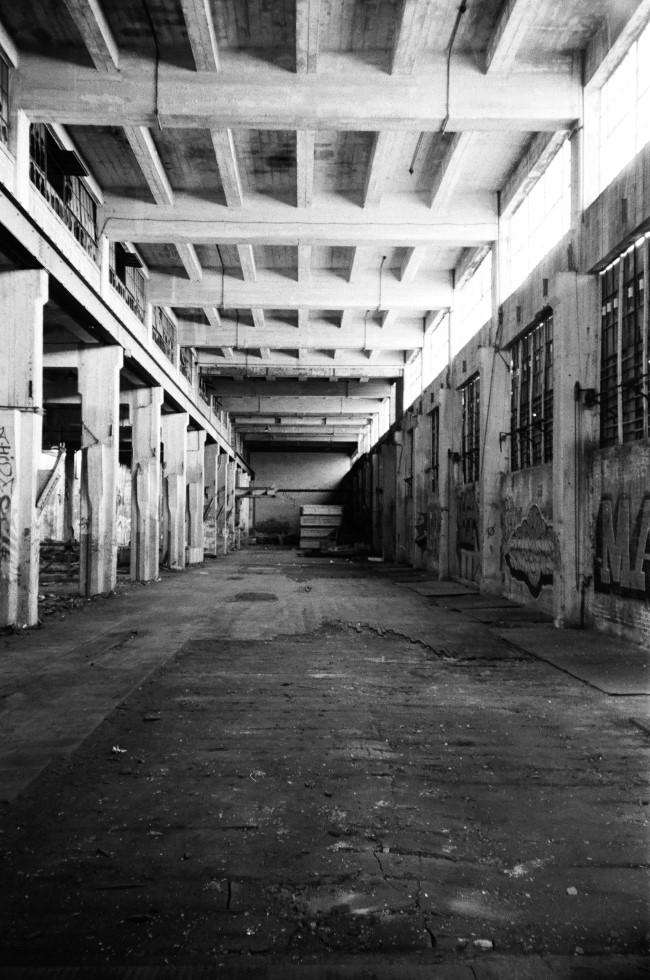 a black and white photo of a cavernous room, lit by windows in the top right. the ceiling is held up by massive concrete pillars on the left.